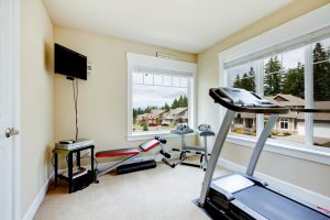 Home gym with equipment, weights and TV with two large windows.